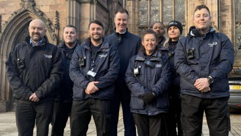 The image shows a group of eight individuals standing together in front of a large, historic stone building with Gothic architectural features. The building has tall, ornate windows with intricate tracery and pointed arches, as well as decorative stone carvings along the roofline. The people are wearing dark uniforms, including jackets and trousers, and several have visible equipment such as radios or body cameras attached to their clothing. The ground is paved with large stone slabs, and part of a vehicle with a yellow license plate is visible on the right side of the image.