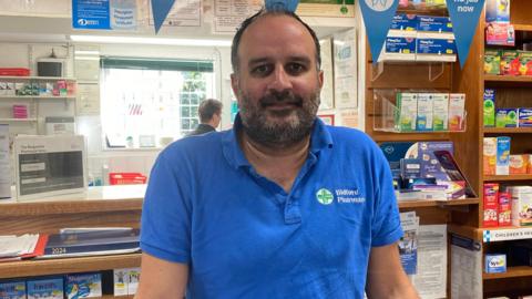 A man with a beard and wearing a blue shirt that says "Bidford Pharmacy" stands behind the counter at a pharmacy.