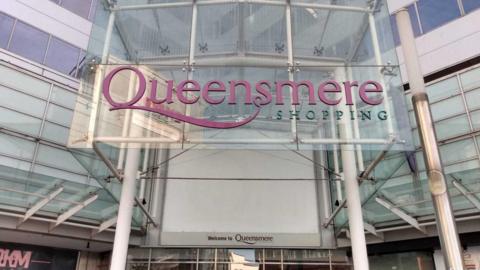 A glass sign above a set of double doors saying "Queensmere shopping" in pink and green writing.