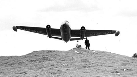 Two men stand on a mound as a large plane flies very closely overhead.