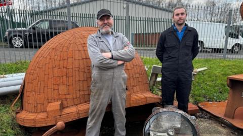 Ray and Sam Lonsdale standing in front of a mining helmet sculpture almost as tall as they are