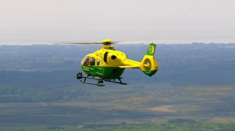 A green and yellow ambulance helicopter is flying mid-air against a backdrop of green land and the coast.