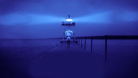 A person walks towards a lighthouse on a dock with an umbrella 