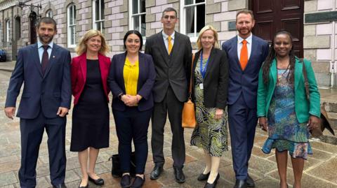 Seven people are standing together outside a large stone-fronted building with tall windows and a dark wooden doorway, posing for a group photograph on a paved street.
The group is dressed in formal business clothing, including suits, jackets and dresses, with several people wearing brightly coloured jackets or ties, suggesting the photo may have been taken at an official or professional event.