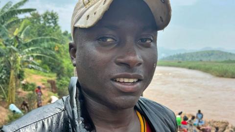 Congolese man Jordan Bita wearing a beige cap and a wet black waistcoat. He is standing on the Burundi side of the River Rusizi - its brown roaring waters can be seen behind him.