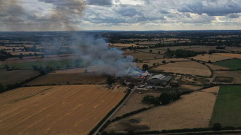 an aerial image of an agricultural farm in the middle of many fields is on fire. the flames are large and orange with grey smoke billowing from them and into the air