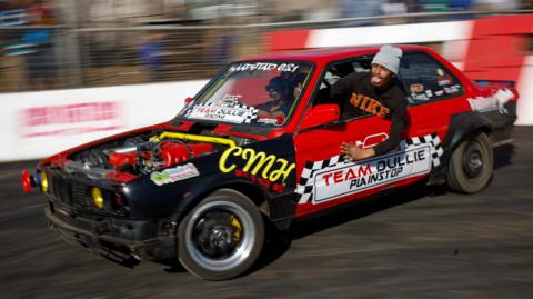 A spinner leans out of a car window to enthrals crowds as he prepares to perform acrobatics from a red and black car while it is moving fast. The car's speed mean that it and the stuntman are in focus but the background is blurred