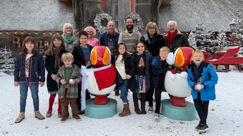 The presenters of television show, the owners of The Book House, and a bundle of children gather around the new look puffins outside the Repair Shop barn in a faux snowy scene. Christmas wreathes and a sledge are in the background.