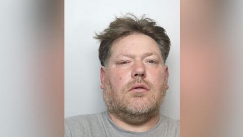 A police mugshot of a man aged around 40, with brown untidy hair and a short beard. He is wearing a grey t-shirt and staring into the camera directly.