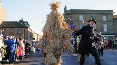 The Straw Bear is paraded through the streets accompanied by attendant keepers, musicians and dancers during the Whittlesea Straw Bear Festival in Whittlesea, Cambridgeshire on 13 January 13 2024. Leepers is leading the Straw Bear by some rope and wears a full black uniform and hat. 