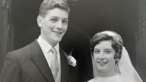 A black and white photo of Anthony and Helen Chandler at their wedding in 1966. Anthony is on the left and has short hair and is wearing a shirt, tie and suit jacket. Helen is on the right and is wearing a white veil and dress.