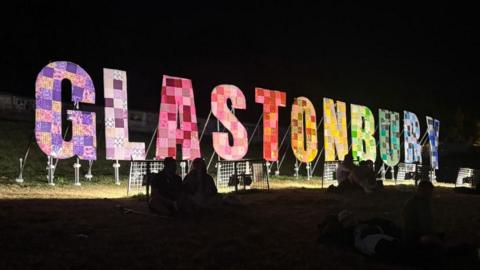 The Glastonbury sign lit up at night. Each letter is a different colour of the rainbow and covered in patchwork. There are people in dark in the foreground. 