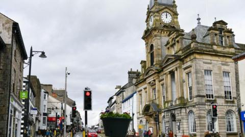 Kendal Town Hall. It is an ornate stone building with a clock tower. Shops and traffic lights can be seen on the opposite side of the street.