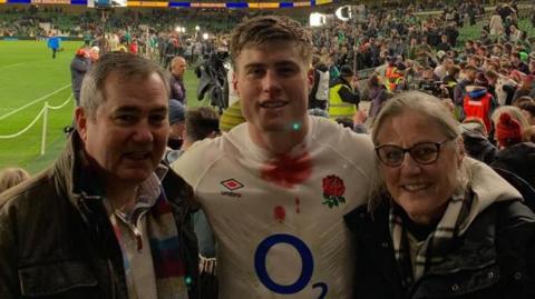 From left-to-right, Cliff Freeman with short grey hair wearing a brown jacket, Tommy Freeman with short dark hair wearing a white England shirt with an O2 logo on the front, and Sara Freeman with short light-coloured hair and glasses wearing a dark jacket with white hood. Tommy has his arms around them and they are standing in a sports stadium with a crowd of people behind them. The rugby pitch is visible to the left.