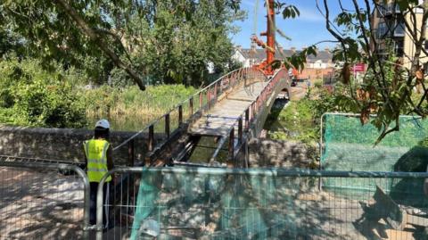 A sunny day with blue sky with a crane in a park area shown lifting a footbridge from the angle of a walker coming up to the bridge. A worker in a green jacket and hard hat is at the front of the bridge which has barriers with green netting on. 