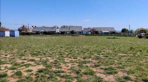 The plot and land in St Dominic in the Tamar Valley. This section is between the Who’d Have Thought It Inn and the village primary school. It has green grass and a few houses can be seen in the background.