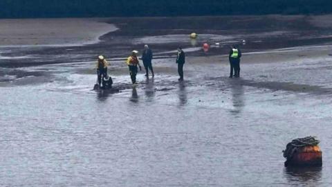 Emergency responders in high‑visibility gear assist a person stuck on a muddy shoreline, using equipment as others look on near the water’s edge.