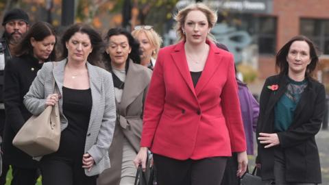 A group of women in a range of ages from mid-30s upwards walking outside a generic British public building.