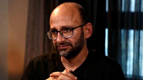 Sami al-Saei during his interview with the BBC. He has a bald head with a short black beard. He wears black-rimmed glasses and a black T-shirt.
