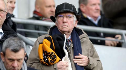 Sir Tom Courtenay, a man in his 80s, with grey hair, is standing in a football terrace with fans around him. he is wearing a black deer-stalker style hat and carrying a black and amber Hull City scarf.