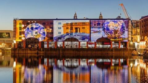 Huge projected images are reflected onto a brick building on the water. The images reflect back into the water in the evening's darkness. The projected pictures show two globes and a picture of a building.