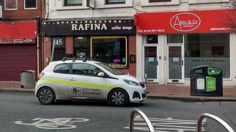 A white car with "CCTV traffic enforcement" written on the side, driving down a street with a row of shops behind it.