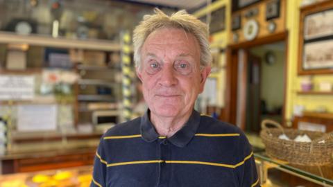 A customer with grey hair and a striped polo top standing in the bakery with pastries behind him