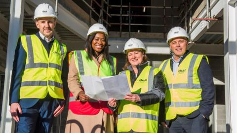 Two men and two women stand outside what looks like a building site. The group are all wearing hi-visibility vests and white hard hats. The two women in the centre of the image are also holding a large sheet of paper.