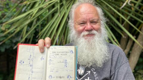 Stephen Mills has grey hair and a long beard. He is wearing a grey T-shirt and smiling while holding the open book, which has drawings of people and written notes on it. He is sitting in front of greenery.