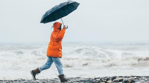 A man wearing a bright orange coat, blue denim jeans and brown boots walks along the seashore. He is holding a navy umbrella.