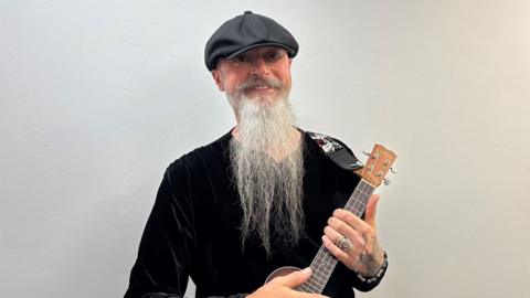 David Danby, a man with a long bear, is wearing a black top and black flat cap. He is holding a ukelele and is standing aganist a white backdrop.