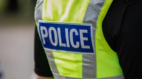 The back of a police officer's yellow high visibility vest with the word POLICE emblazened across it on a blue rectangle.