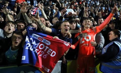 Coventry City fans at the front of the Ewood Park stands, hold a Promoted 2025/26 banner while hugging Sky Blues player Josh Eccles who has a huge grin on his face with an arm aloft