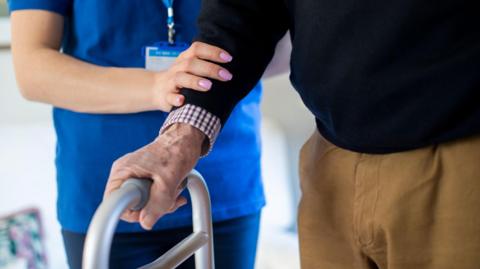 Close Up Of Senior Man With Hands On Walking Frame Being Helped By Care Worker.