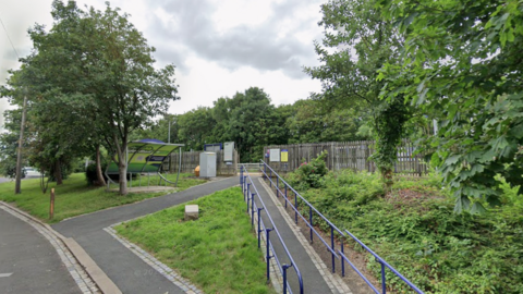 The outside of a deserted Seaton Carew station with two ramps leading up to a platform where a gap in a wooden fence allows access to the station. One of the ramps, which are not very steep, has blue/purple hand rails on each side. There are several trees in full leaf and a bike shelter. There are clouds in the sky on a grey day. 