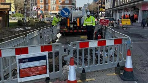 Men in high-visibility jackets working behind a grey and red plastic barrier. There are cones and barriers alongside road closure signs. 