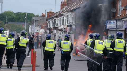 A line of police officers facing a burning car on a street of terraced houses and shops. A large group of people is in the distance