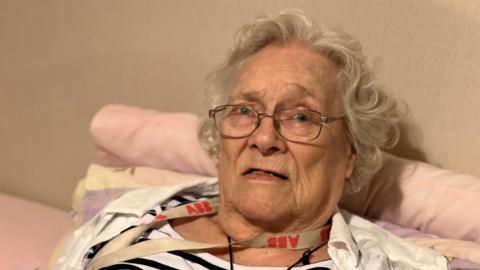 Henrietta is sitting up in bed against pink pillows and bedding. She has got curly white hair and is wearing metal framed glasses. She is wearing a black and white stripey nightie and a white shirt and has 2 lanyards around her neck.