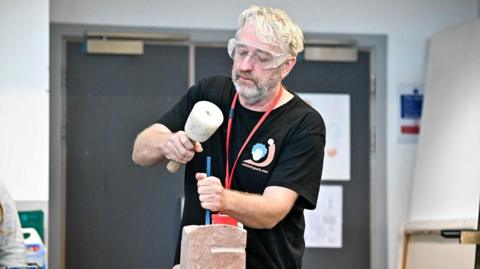 Rob MacDonald working on a stone sculpture. He is wearing a black t-shirt, protective glasses and a red lanyard. He has white hair and beard. There is a grey door behind him.