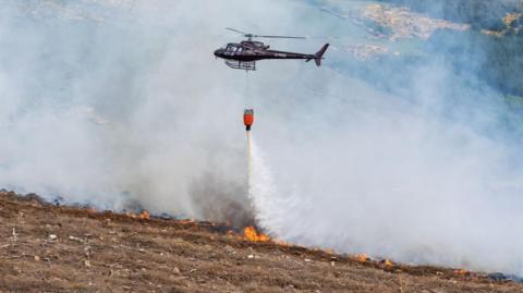 A helicopter waterbombs flames in a wildfire on a barren hillside. Water pours from a large red bucket under the helicopter.