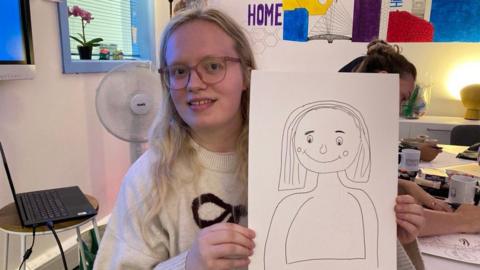 Hannah holding a picture she drew of herself. The drawing is in black pen on white A4 paper and shows a woman with her hair down and she is smiling. 