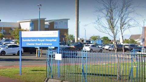 A blue sign reading Sunderland Royal Hospital Chester Road Entrance. There is a car park behind it, with the hospital's tall beige and grey buildings further back. A blue fence is separating the grassland near the car park from the street pavement. 