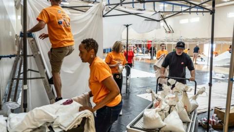 Volunteers in orange and black t-shirts build a blanket fort while a man pushes a trolled with white bags on it. 