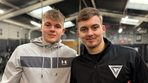 A man with blonde hair wearing a grey jacket, which has a dark grey stripe, stands next to his brother who is wearing a black and red jacket from the show Gladiators