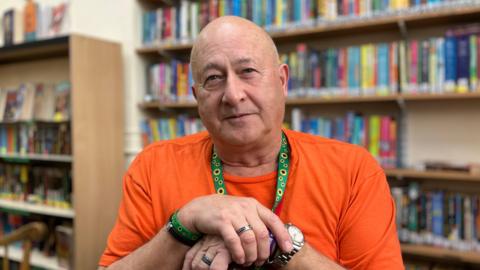 John Hyde sitting in the school's library with his wrists crossed over each other. He is wearing a bright orange t-shirt and a green lanyard with sunflowers on, with a matching wristband. He is wearing a large silver watch and silver rings. He is bald and is looking directly into the camera with a slight smile. 