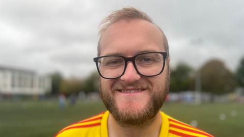 Aaron Richards in yellow and red football shirt, smiling at the camera. The background is blurred, but appears to be a football pitch. 