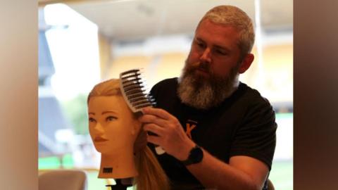 Simon is practicing on a hair model but using a large brush the wrong way round as he tends to the hair. He has a black t-shirt on and a long grey beard.