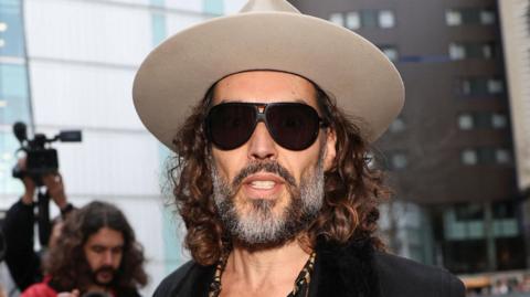 Russell Brand arriving at Southwark Crown Court wearing a light grey fedora hat, dark glasses, a leopard print shirt unbuttoned halfway down his chest, a cross on a necklace, and a dark jacket
