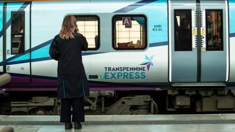 A woman stands on a train platform facing a TransPennine Express train.  She has her back facing the camera and is dressed in black.