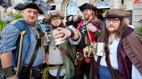 Four men dressed in pirate costumes. They are all looking at the camera and posing, holding drinks and other props.